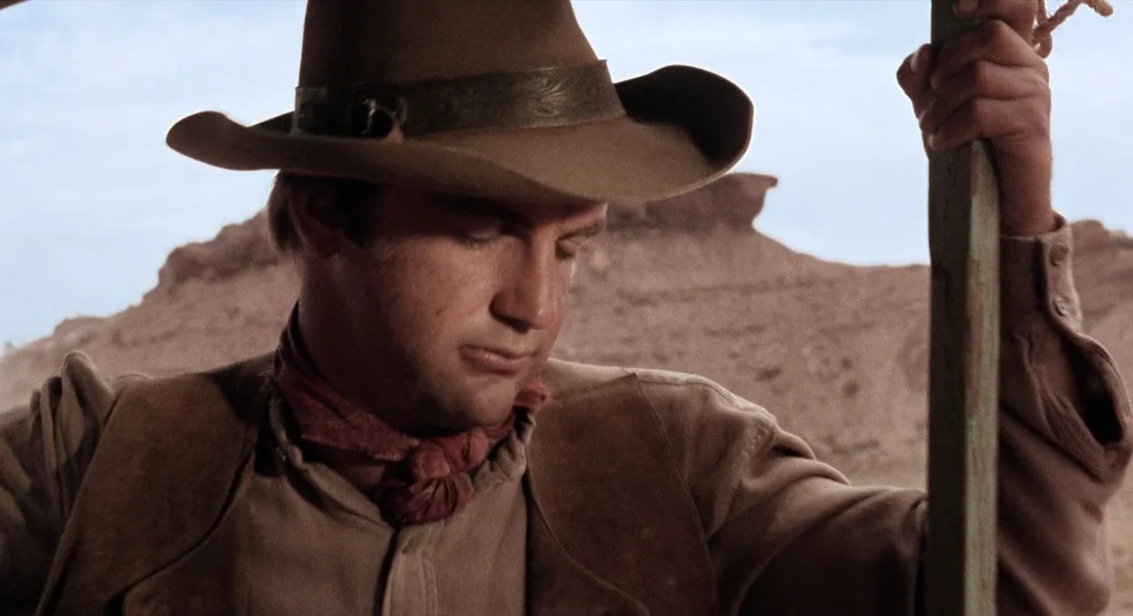 Ride In The Whirlwind (1966) movie still: medium — A man in western attire stands in a vast, arid desert environment. He grips a wooden post while…