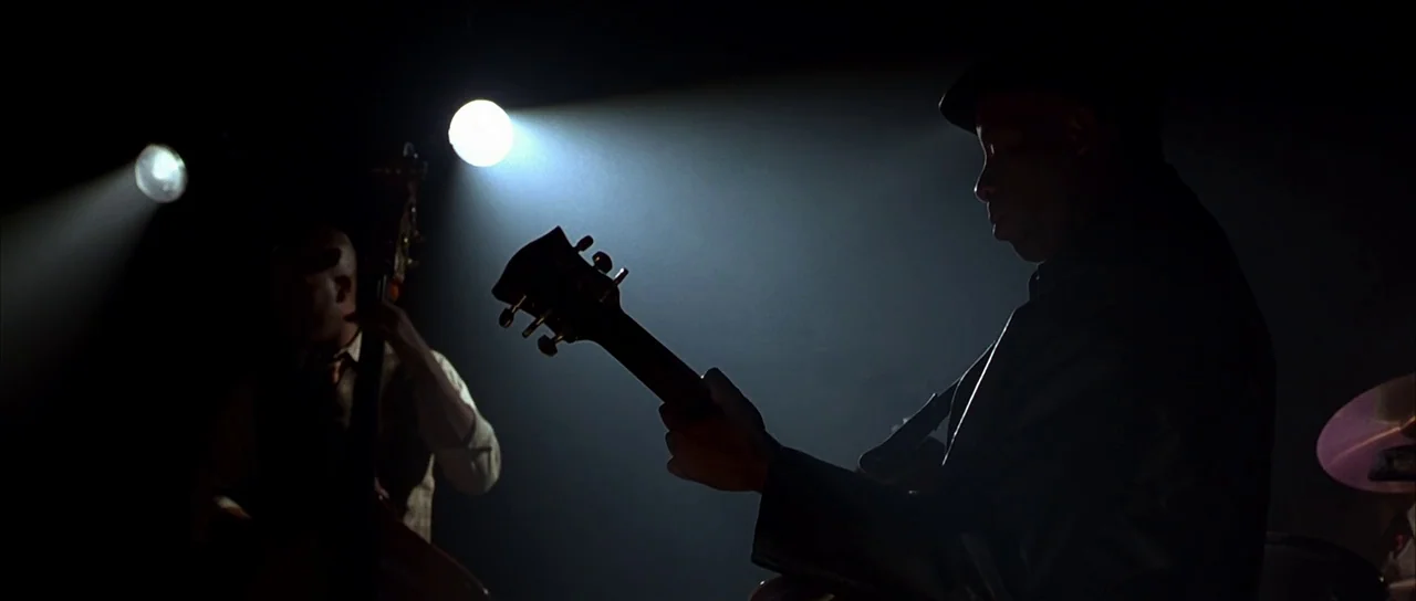 Dark City (1998) movie still: medium — Two musicians perform on a darkened stage illuminated only by isolated overhead spotlights. A guitar player…