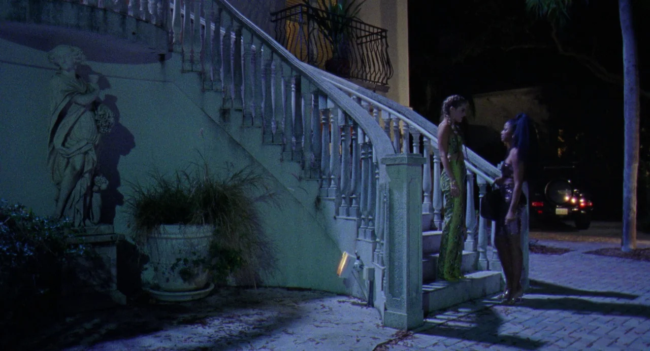 Zola (2020) movie still: wide — A stone outdoor staircase with a classic statue stands next to a driveway at night. Two young women stand on the…