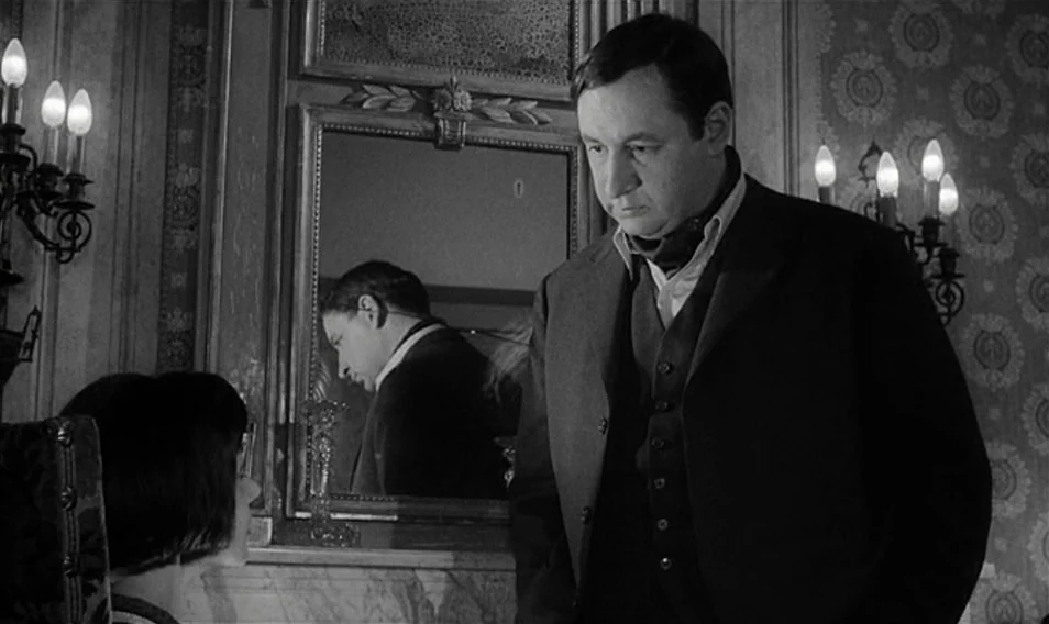 Thérèse Desqueyroux (1962) movie still: medium — An ornate drawing room features patterned wallpaper and a large central mirror over a mantle. A man…