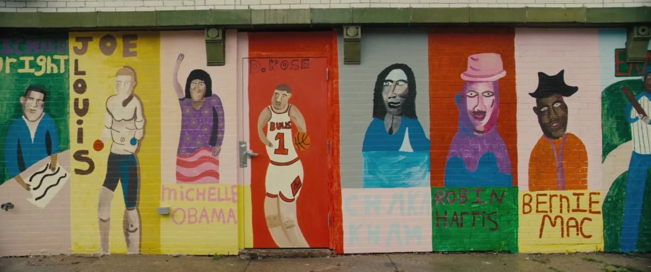 Chi-raq (2015) movie still: wide — A brick wall features a brightly painted mural portraying various notable cultural figures. The figures are…