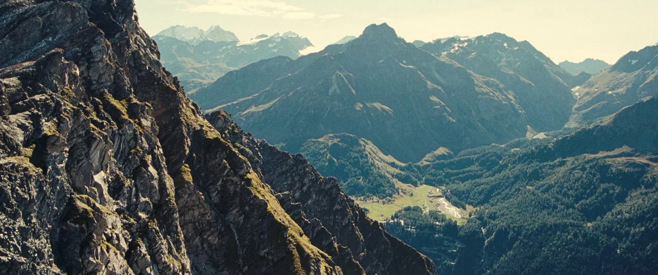 Clouds of Sils Maria (2014) movie still: establishing — A sprawling mountain range features rugged craggy peaks towering over a dense evergreen…