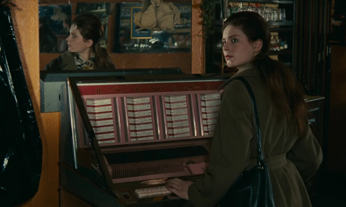 Muriel (1963) movie still: medium — A dimly lit tavern featuring a vintage jukebox and a mirror on an orange wall. A woman with long hair wearing an…