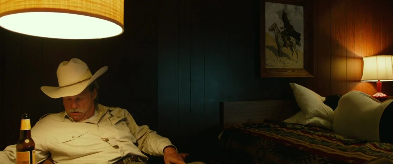 Hell or High Water (2016) movie still: medium — A dark wood-paneled room contains a bed and a man in a cowboy hat. The man sits in the foreground…