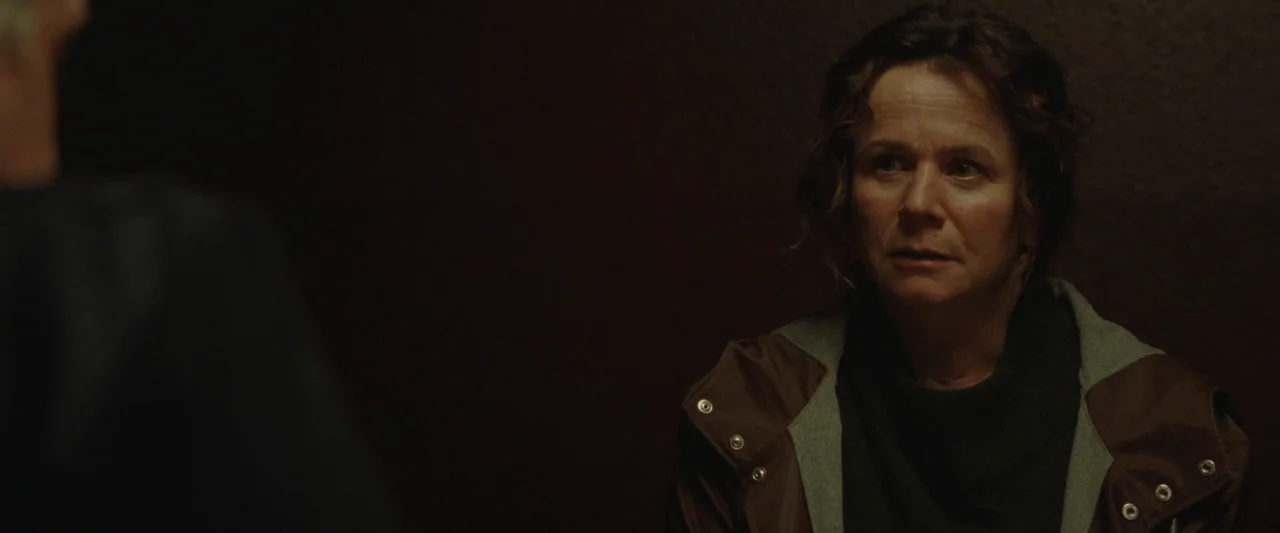 God’s Creatures (2022) movie still: medium — A dark, dimly lit interior environment with no visible architectural details. A woman in a brown…