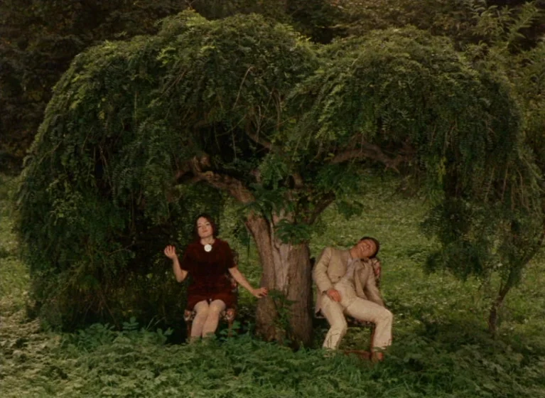 Fruit of Paradise (1970) movie still: wide — A man and woman sit in chairs placed under a large weeping tree outdoors. The scene is set in a green…
