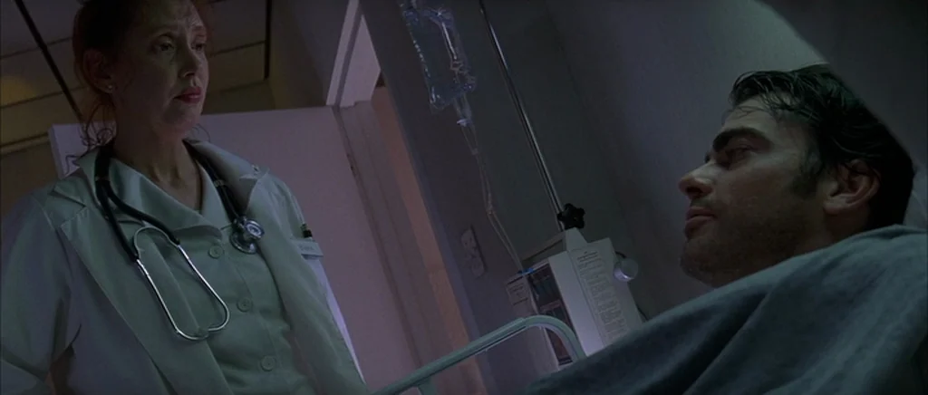 The Underneath (1995) movie still: medium — A dimly lit hospital room features a medical patient bed and IV stand. A female nurse in a white coat…