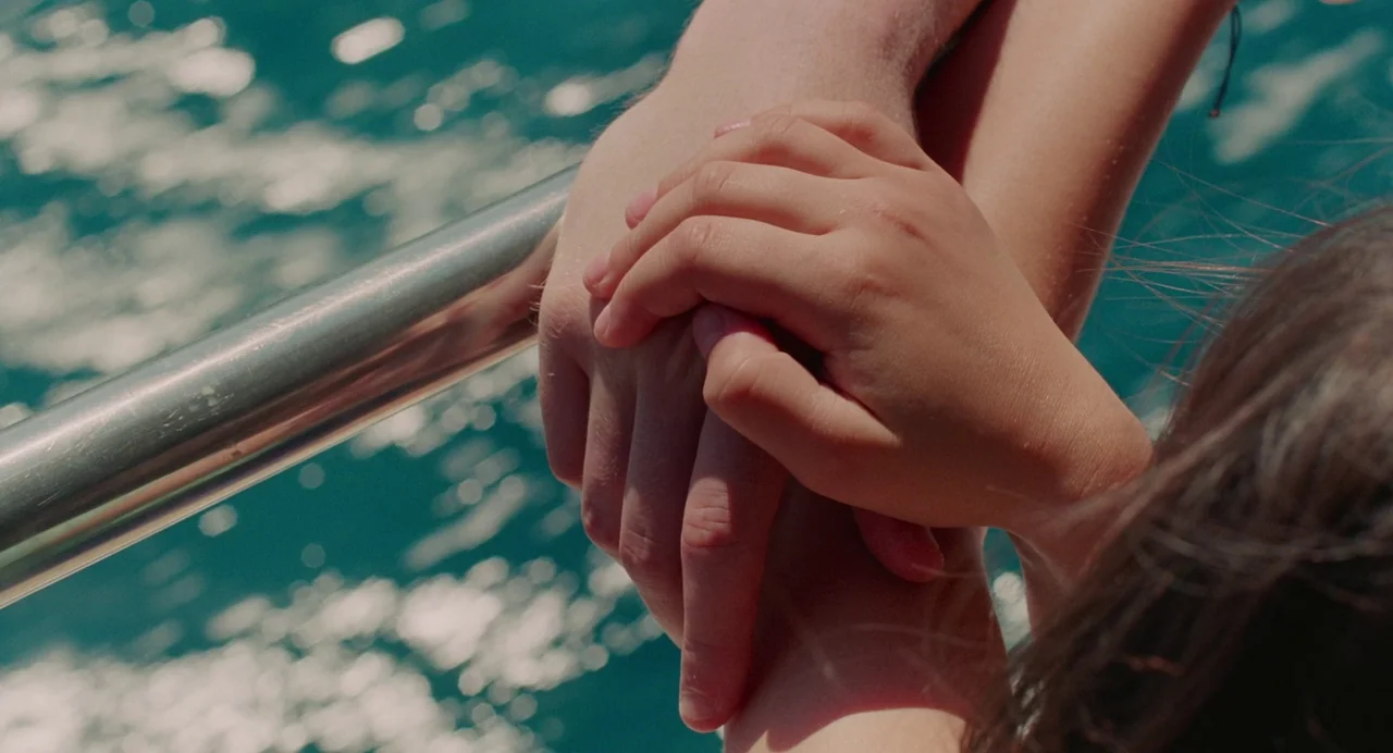 Aftersun (2022) movie still: closeup — A metal railing is visible over blue water with sunlight reflecting on the surface. Two hands rest on the…