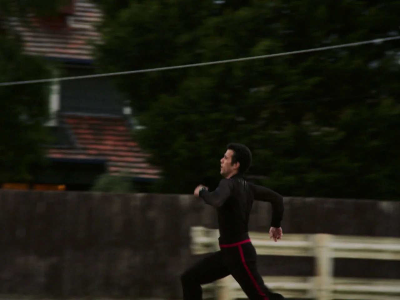 Of An Age (2022) movie still: fullbody — A man runs past a residential fence and green trees during twilight. The background shows a dark house with…