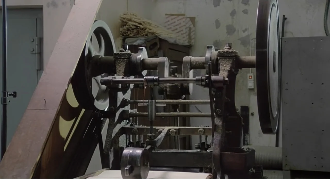 The Match Factory Girl (1990) movie still: medium — A cluttered workshop contains a heavy, aged industrial machine with large metallic flywheels.…