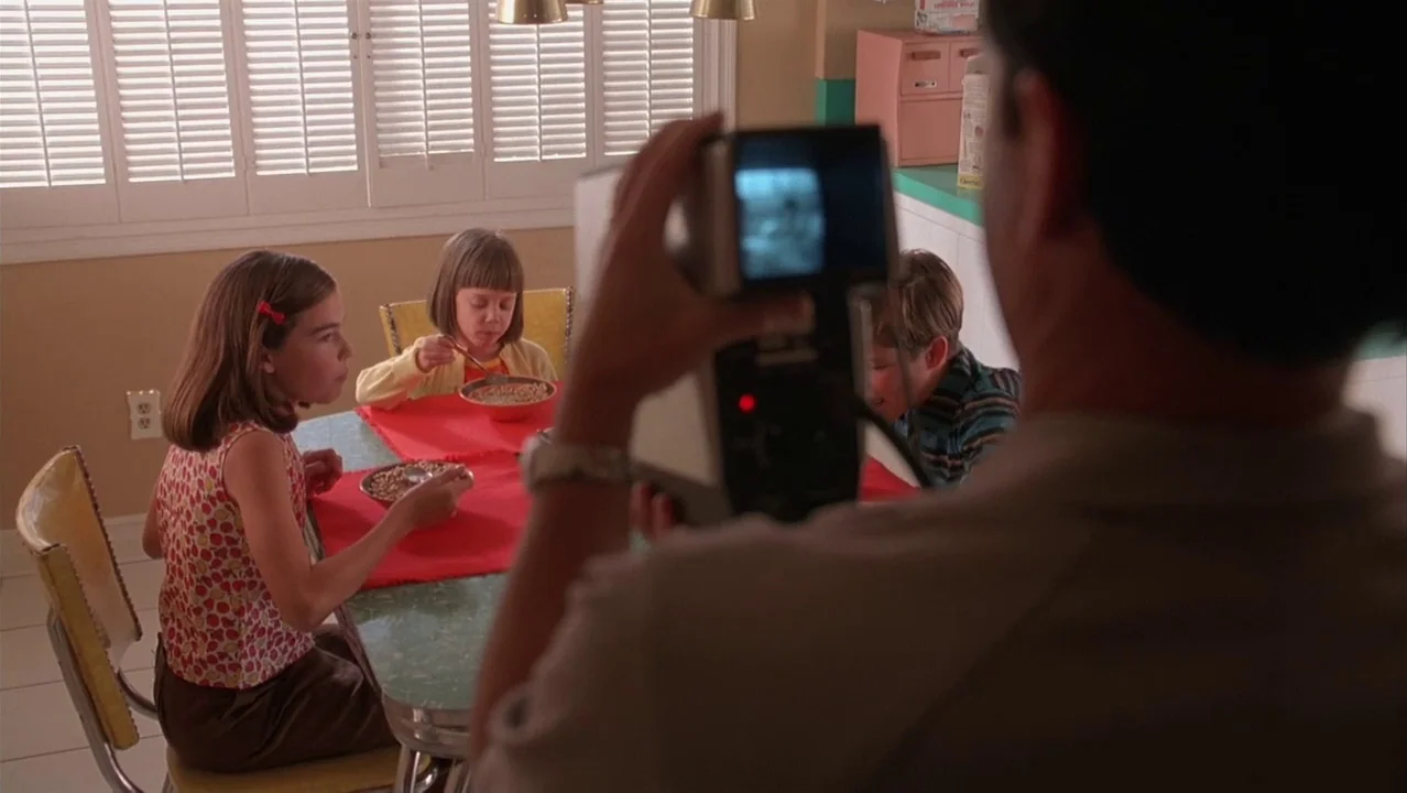 Auto Focus movie still: medium — A 1990s-style room features yellow walls, louvered window shutters, and a green laminate table. A person holds a…