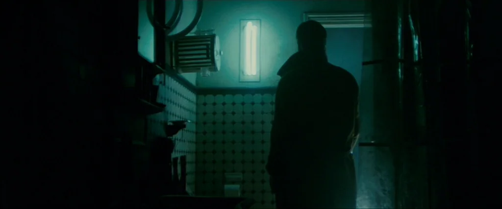 Blade Runner (1982) movie still: medium — A dimly lit room with small patterned tiles on the walls beneath a light. A person in a dark coat stands…