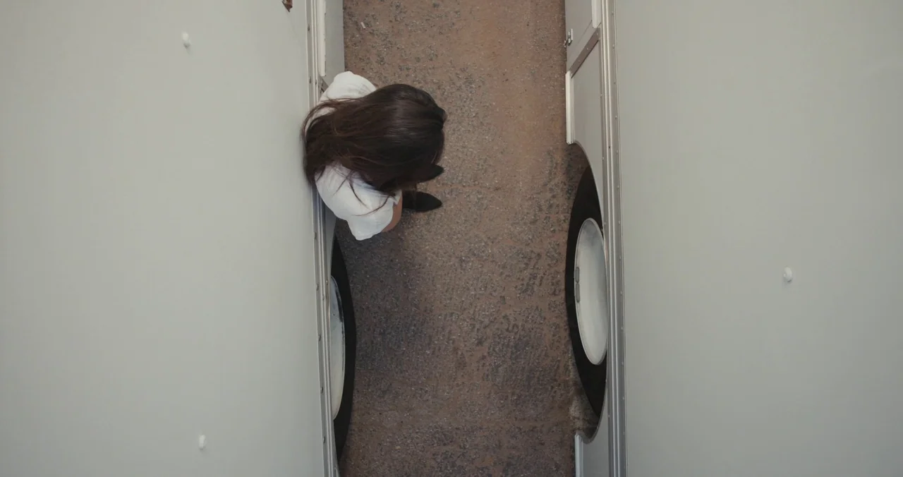 Violet (2021) (2021) movie still: wide — A narrow space between two white trucks parked on a weathered asphalt surface. A person with dark hair…