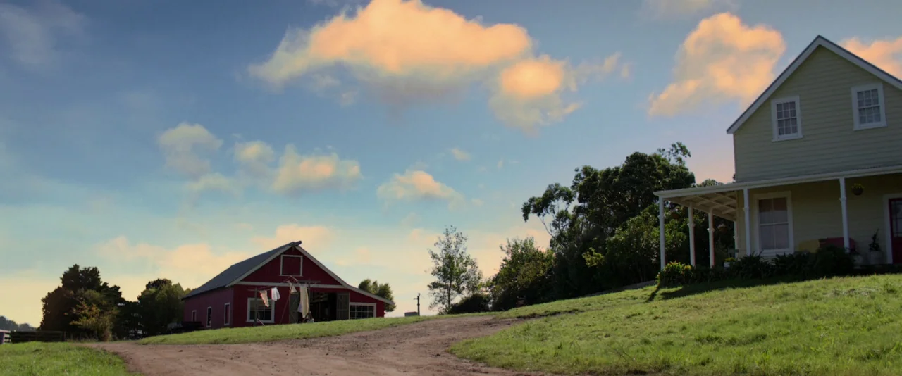Pearl (2022) movie still: establishing — A rural setting features a small red barn and a yellow house on grass. A dirt path leads between the two…