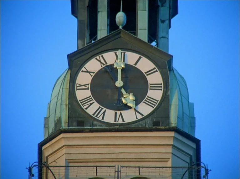 Fox and His Friends (1975) movie still: closeup — A stone clock tower with a prominent Roman numeral dial sits against a blue sky. The clock hands…