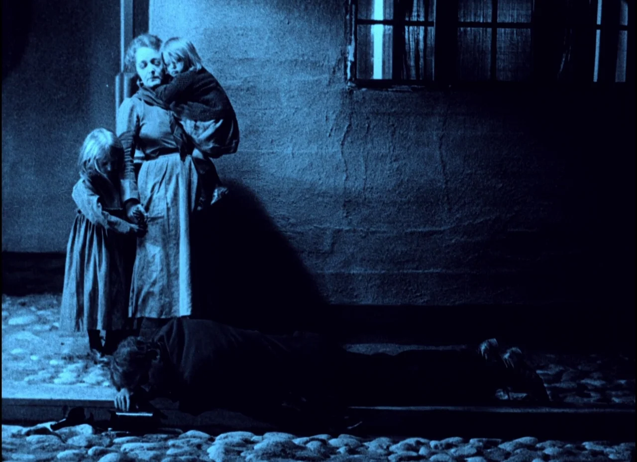 The Phantom Carriage (1921) movie still: fullbody — A dark cobblestone street at night is illuminated by a single window light. A mother holds one…
