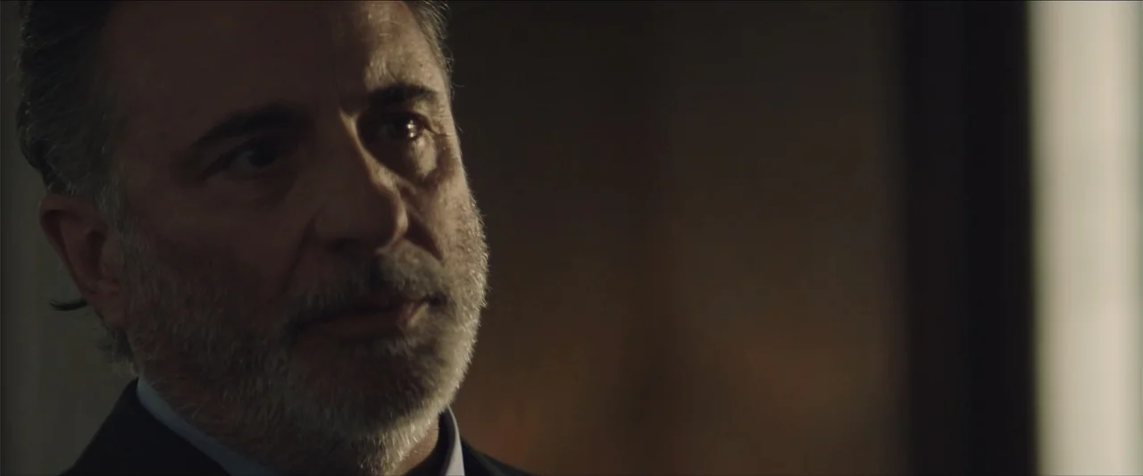 Redemption Day (2021) movie still: closeup — A dimly lit room with shadows obscuring the background. An older man in a suit looks off-screen.