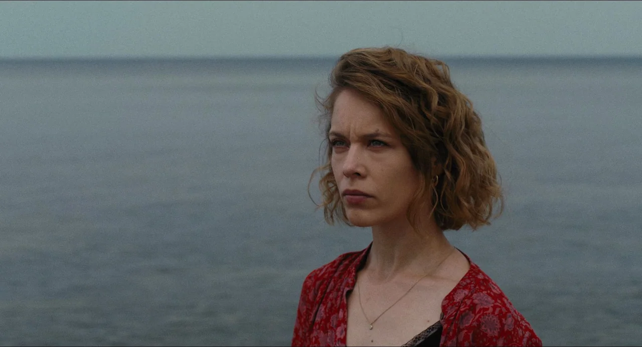 Afire (2023) movie still: medium — A woman stands before a vast, calm sea under an overcast grey sky. She gazes off-camera while wearing a patterned…