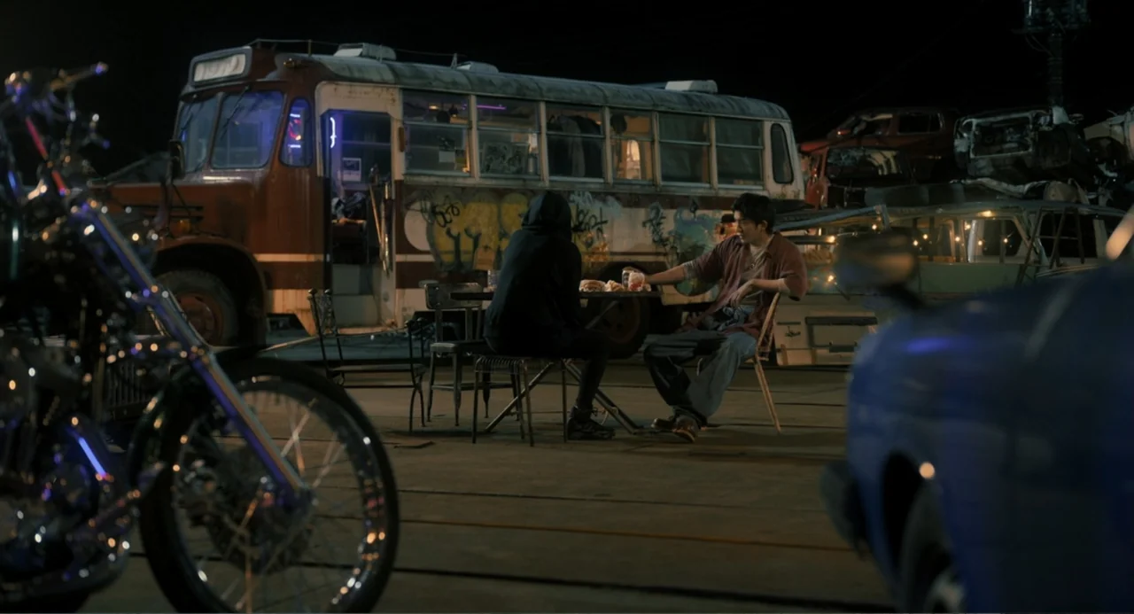 Knuckle Girl (2023) movie still: medium — An outdoor junkyard at night contains a weathered bus covered in graffiti. Two people sit at a small table…