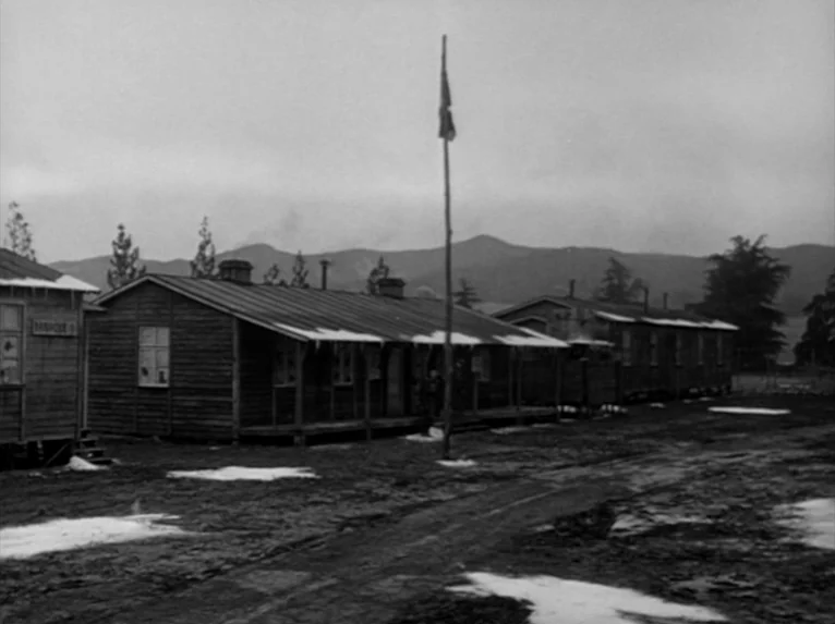Stalag 17 (1953) movie still: wide — A cluster of weathered wooden barracks buildings stands on a muddy, partially frozen field. A tall flag pole…