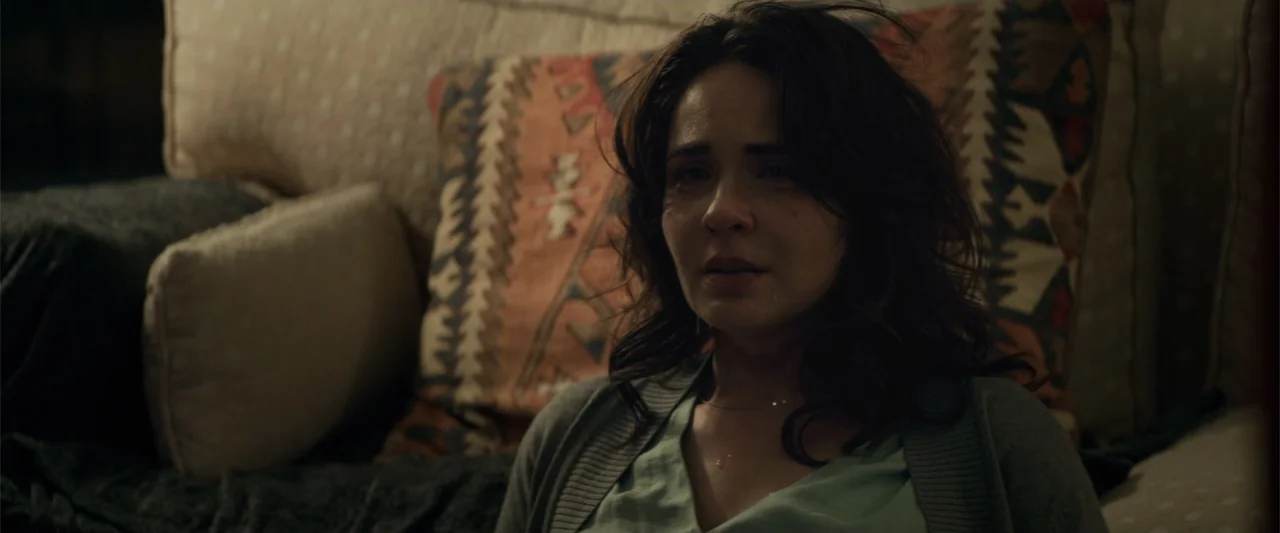 Human Animals (2020) movie still: closeup — A dimly lit room contains a patterned cushion on a sofa. A woman sits on the couch with a tear-streaked…