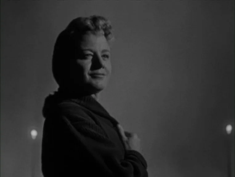 The Night Of The Hunter (1955) movie still: medium — A woman stands against a dark, featureless night background with two distant light sources. She…
