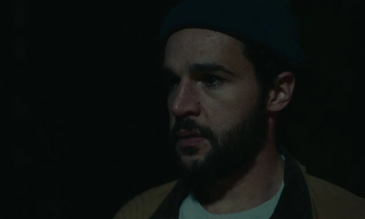 Black Bear (2020) movie still: closeup — A man with a beard sits against an unlit, dark background. He wears a beanie and a light-colored shirt…