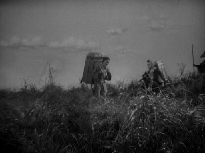 The Men Who Tread on the Tigers Tail (1945) movie still: wide — An open grassy field stretches toward a clouded horizon under an overcast sky. Two…
