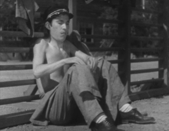 Tokyo Chorus (1931) movie still: fullbody — A man sits on the ground against a wooden fence in a dusty area. He is shirtless and wears a peaked cap…