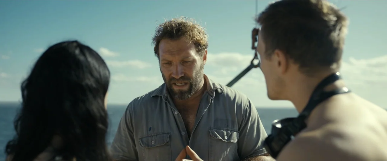 Dangerous Animals (2025) movie still: medium — A wooden boat deck under a clear blue sky overlooks an open sea horizon. A man with a curly beard…