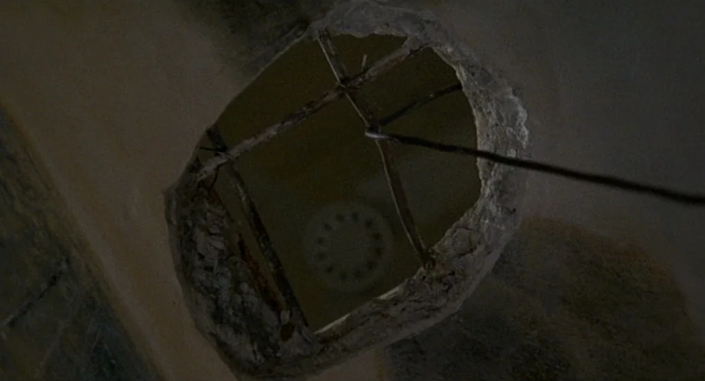 Furia (1999) movie still: closeup — A dark, circular hole is broken through a rough plaster or concrete ceiling. Metal reinforcement bars cross the…