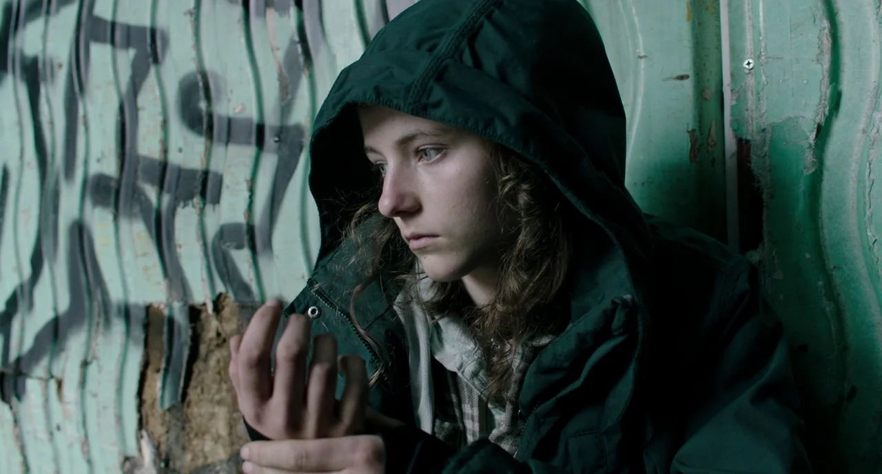 Leave No Trace (2018) movie still: closeup — A person sits in front of a textured teal wall covered in black graffiti. They are looking down at…