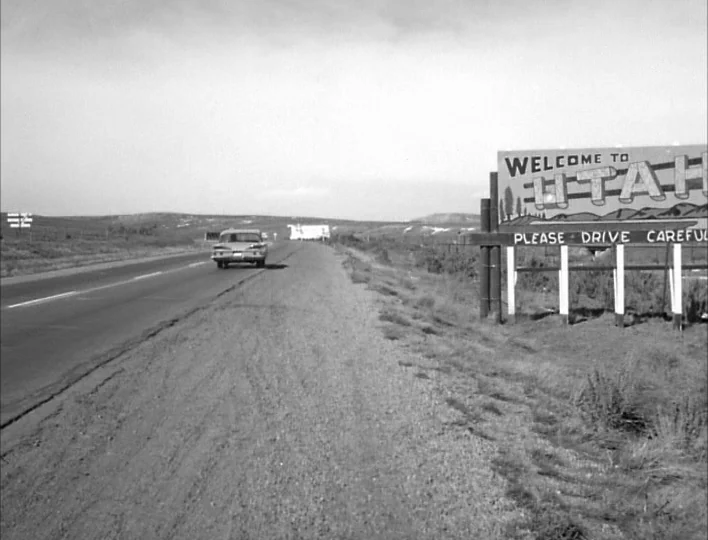 Carnival of Souls (1962) movie still: wide — A desolate desert highway features a large welcome sign on the dusty roadside. A single vintage sedan…