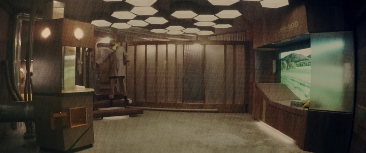 Domain (2017) movie still: wide — A utilitarian room features wood-paneled walls and a grid of overhead hexagonal lights. A digital screen displays…