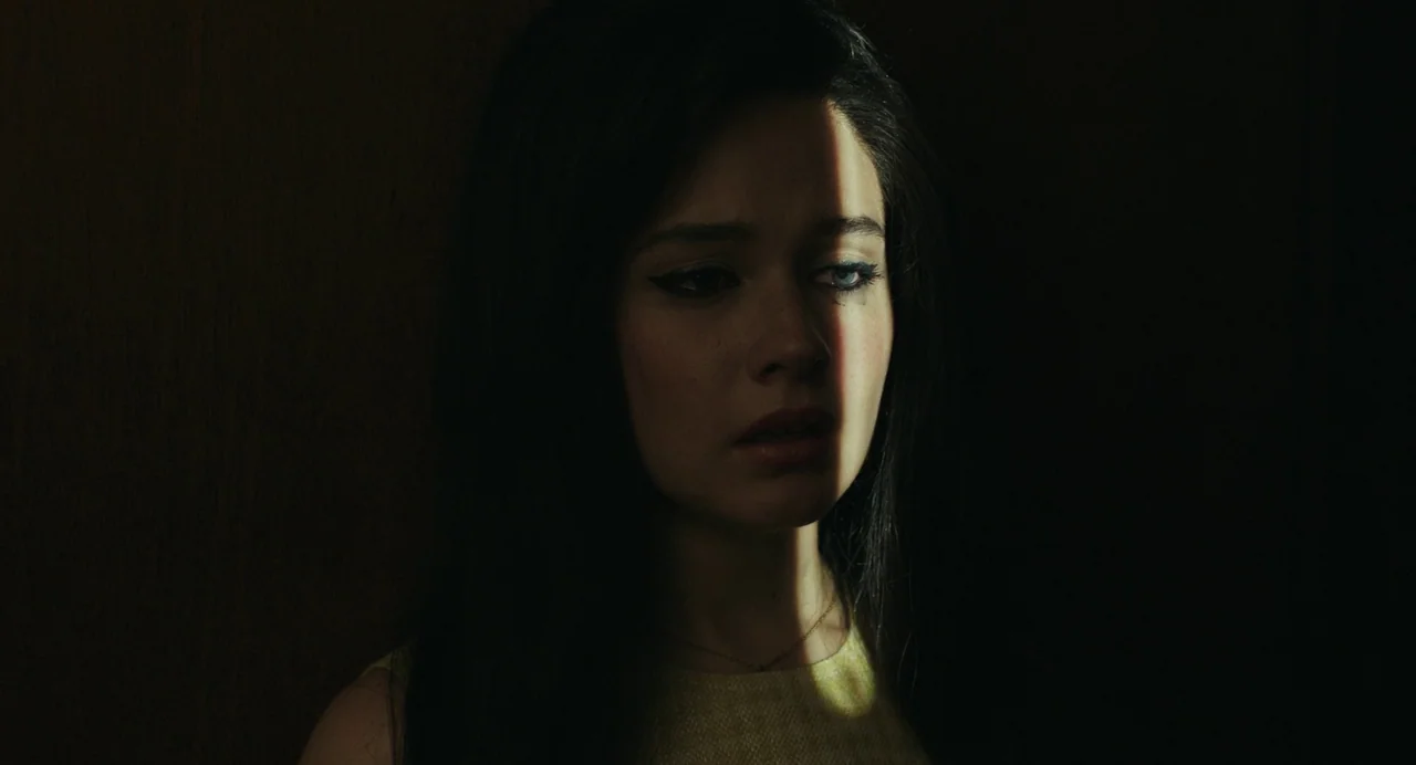 Priscilla (2023) movie still: closeup — A person stands in a dimly lit space with a singular light beam. A woman is partially obscured by shadows…