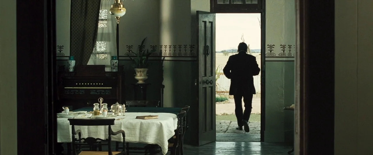 The Proposition (2005) movie still: wide — A rustic dining room features a laid table and a patterned wallpaper border. A man in a dark suit walks…