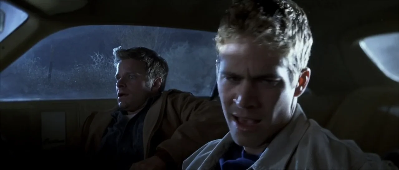 Joy Ride (2001) movie still: medium — The interior of a car at night with darkness visible through the windows. Two men sit side-by-side in the…