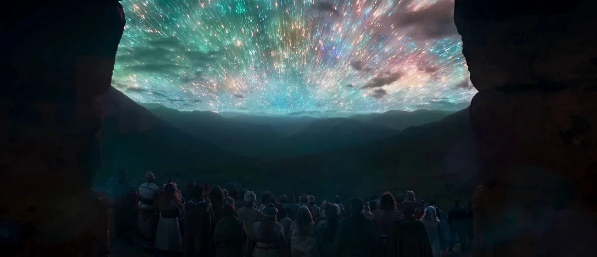 Andor (2022) movie still: wide — A dense crowd of people in rustic attire stands at the edge of a cave. They look out at a vast mountain range under…