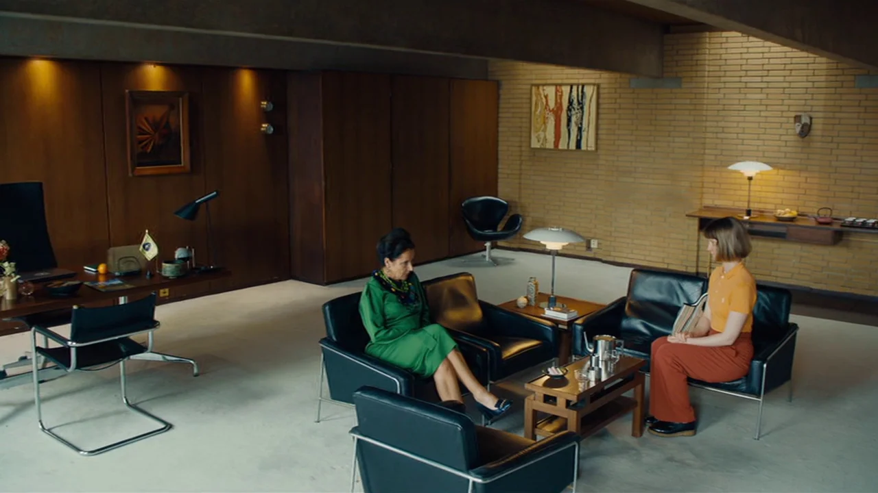 Club Zero (2023) movie still: wide — A spacious room features mid-century modern furniture, wood-paneled walls, and yellow brick accents. Two women…