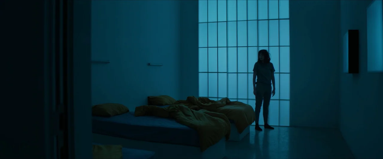 Double Blind (2023) movie still: wide — A austere room features two small beds and a large, glowing rectangular window panel. A young woman in…