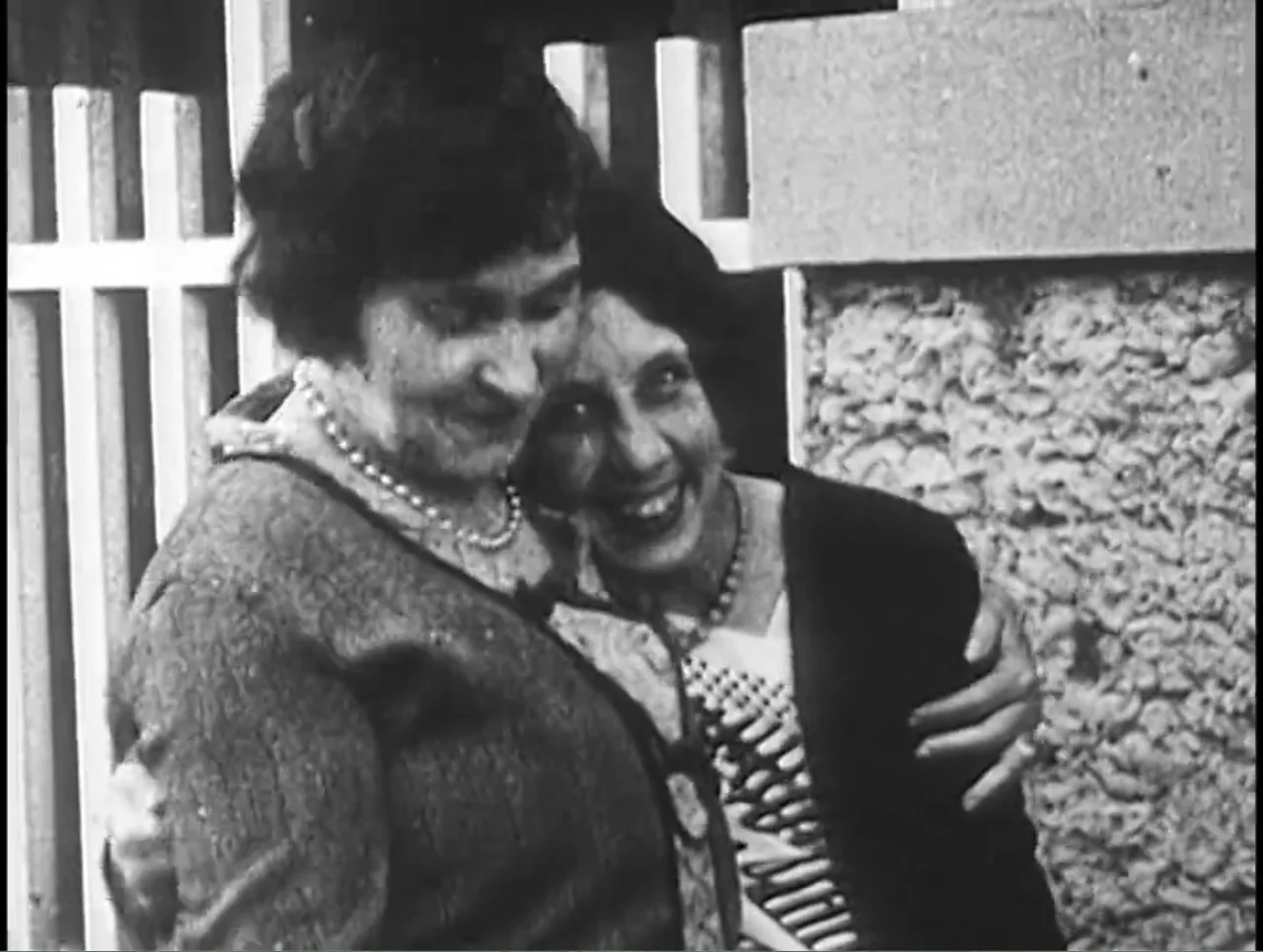 First Cousin Once Removed (2012) movie still: medium — Two women embrace against a rough stone wall with a white picket fence nearby. They smile at…