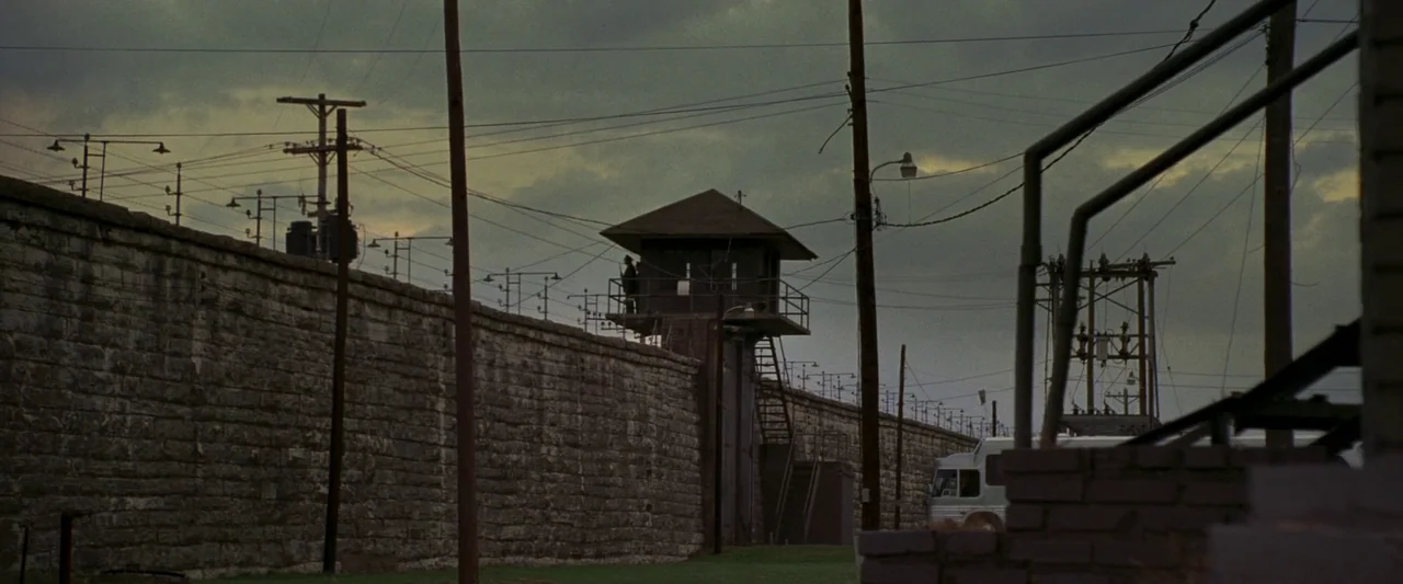 Walk The Line (2005) movie still: wide — A long stone prison wall stretches across the frame with a central guard tower. Overhead power lines…