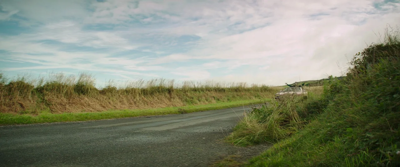 Mindhorn (2016) movie still: wide — A quiet rural road is bordered by grassy banks and tall wild grasses. A small light-colored car is visible in…
