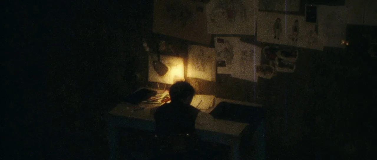 The Orphanage (2007) movie still: wide — A dimly lit workspace with drawings taped to the wall behind a desk. A young child sits at the table,…