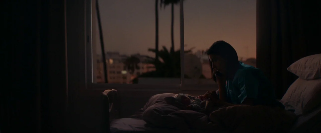 Animalia (2023) movie still: medium — A dark hospital room at dusk overlooks a city skyline with palm trees. A woman sits on the side of a bed…