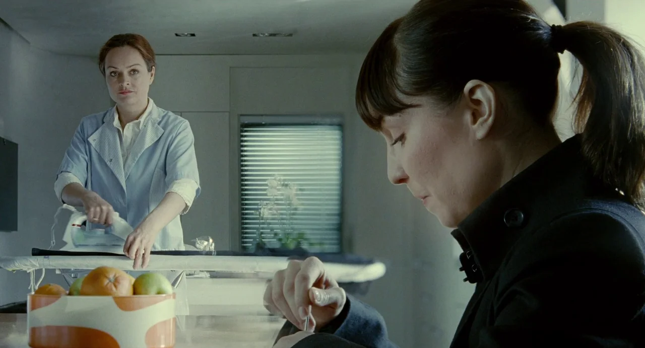 Passion (2012) movie still: medium — A clean modern room features a woman ironing clothes on a board. Another woman sits at a table in the…