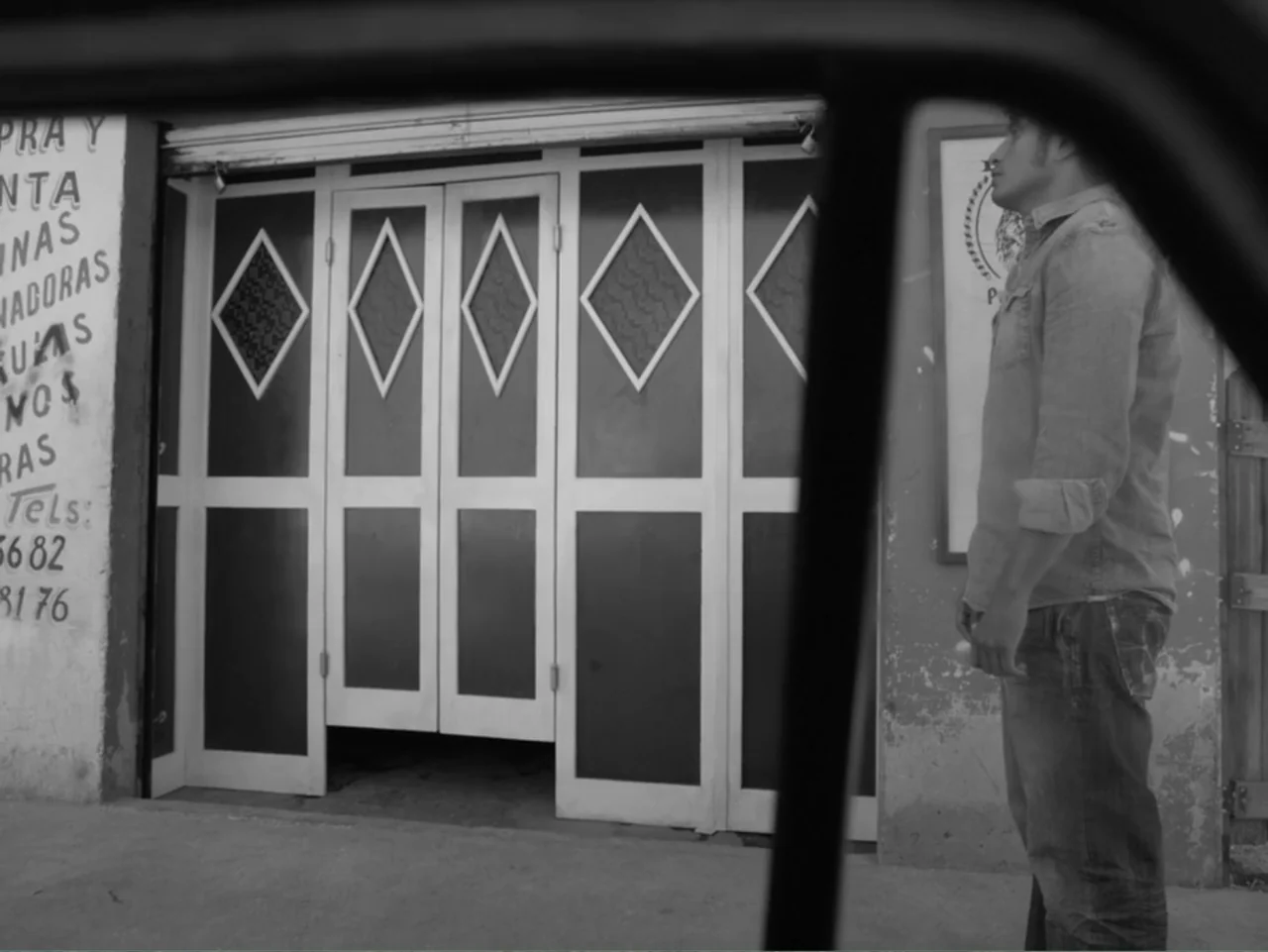 Güeros (2014) movie still: medium — A sidewalk exterior features a white folding door with diamond-shaped cutouts. A man stands in profile before…
