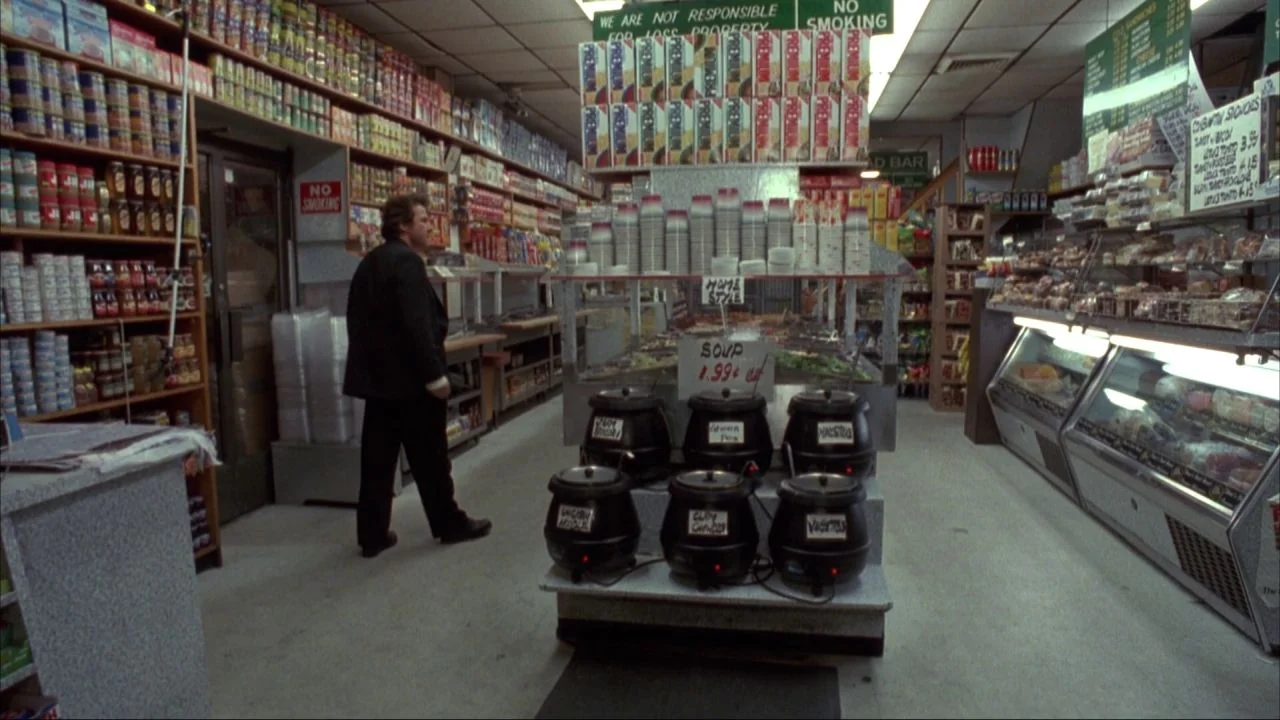 Bad Lieutenant (1992) movie still: fullbody — A cramped food market with floor-to-ceiling shelves packed with canned goods and snacks. A man in a…