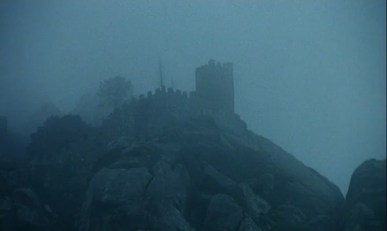The Rose King (1986) movie still: establishing — A stone castle keep and wall perimeter sit on a jagged, rocky cliffside. Heavy blue fog obscures…