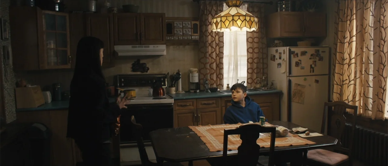 All Fun and Games (2023) movie still: medium — A residential kitchen features wood cabinetry, a dining table with patterned cloth, and appliances. A…